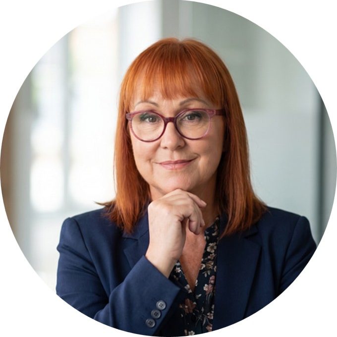 Andrea Kennedy A red haired woman with glasses and hand place under her chin, smiling at the camera.